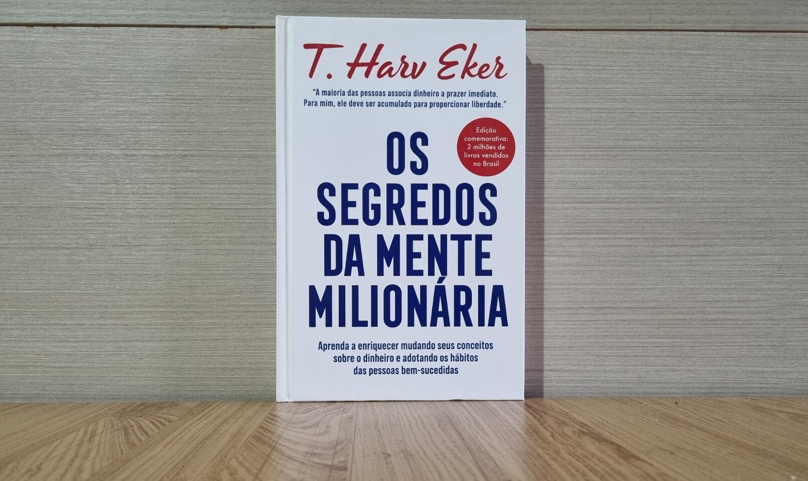 Foto real da capa do livro 'Os Segredos da Mente Milionária' de T. Harv Eker, apoiado sobre uma mesa de madeira.