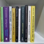 Foto real de doze livros de finanças e investimentos alinhados lado a lado sobre uma prateleira.