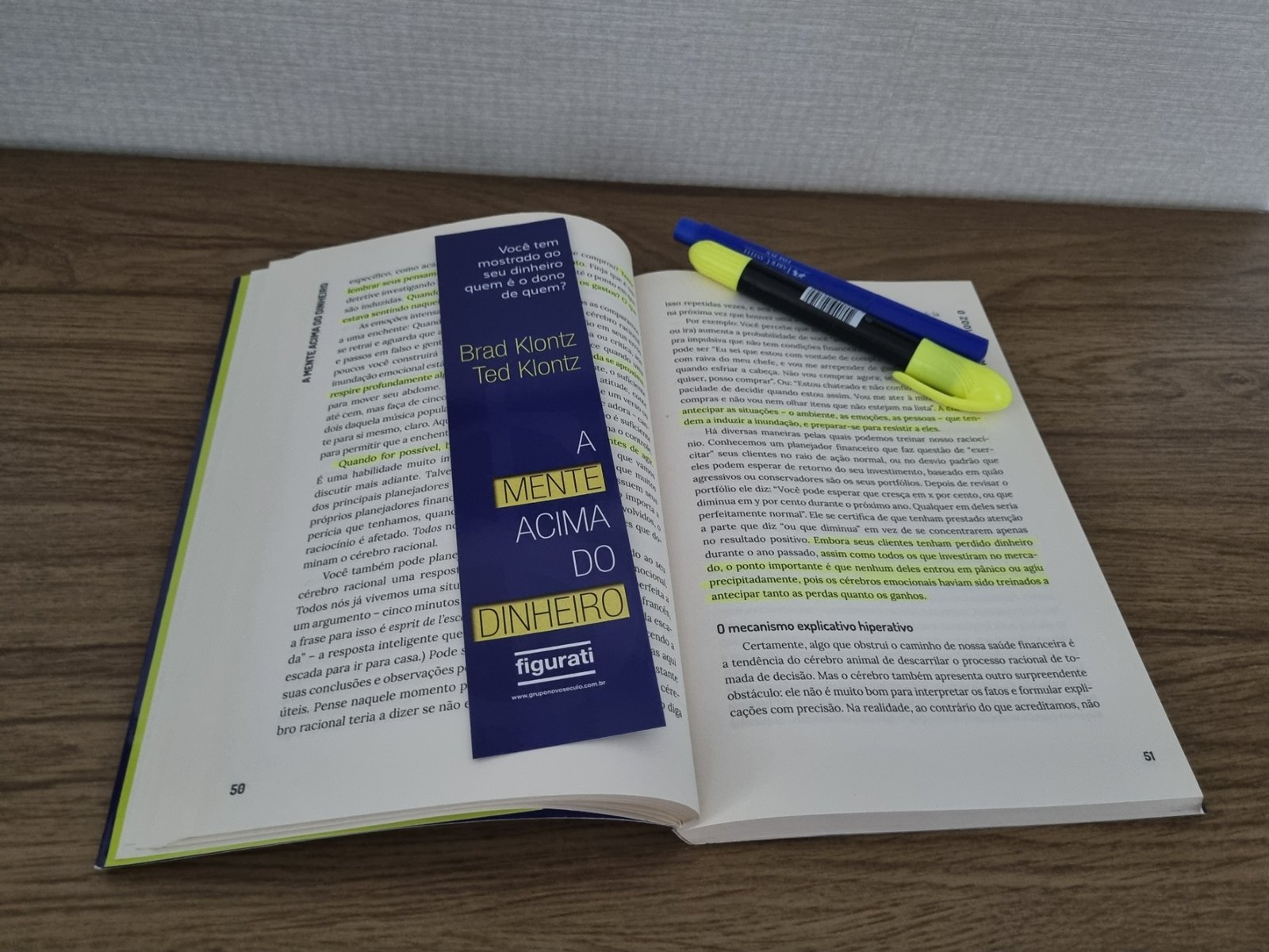 Livro A Mente Acima do Dinheiro aberto sobre uma mesa de madeira, mostrando páginas com trechos destacados em caneta marca-texto amarela, acompanhado de marcador de páginas personalizado.