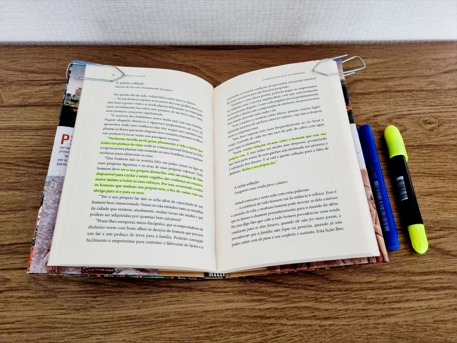Livro O Homem Mais Rico da Babilônia aberto sobre uma mesa de madeira, mostrando páginas com trechos grifados em verde e duas canetas marca texto ao lado.