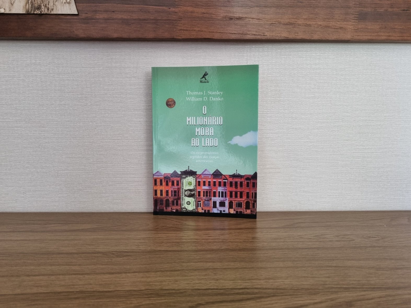Foto autoral da capa do livro O Milionário Mora ao Lado de Thomas J. Stanley e William D. Danko sobre uma bancada de madeira.