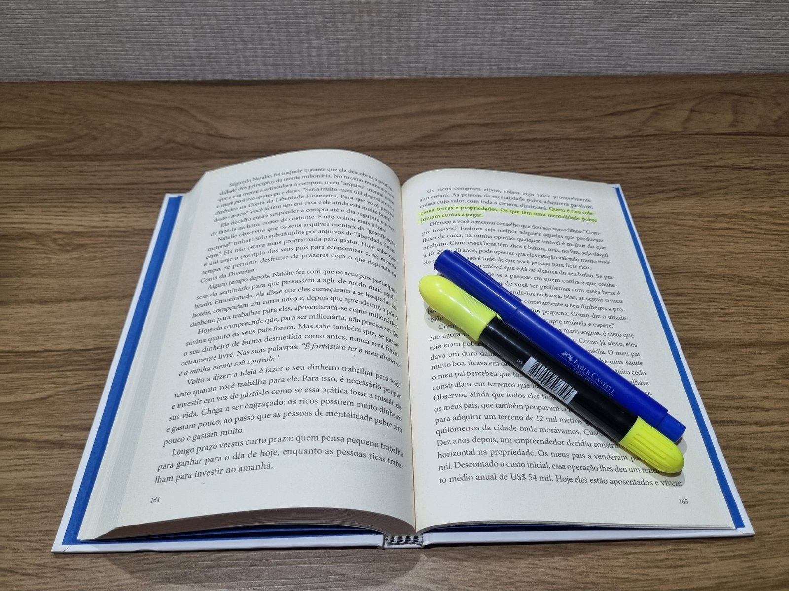 Livro Os Segredos da Mente Milionária aberto sobre uma mesa de madeira, com páginas grifadas em verde e uma caneta marca-texto amarela apoiada sobre elas.