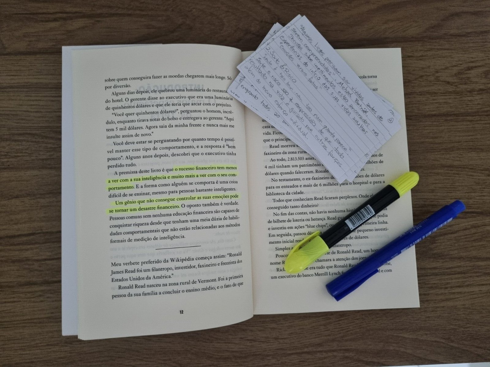 Livro A Psicologia Financeira aberto sobre uma mesa de madeira, mostrando páginas com trechos destacados em caneta marca-texto amarela, acompanhado de canetas e papéis de anotação.