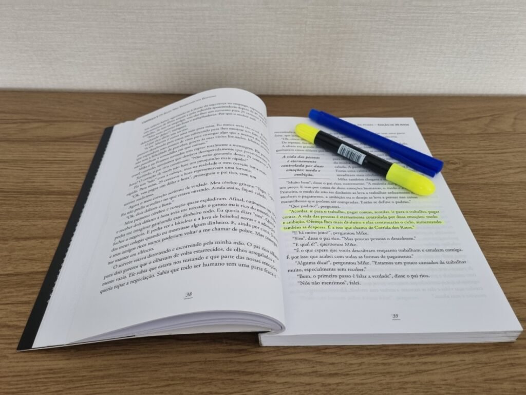 Livro Pai Rico, Pai Pobre aberto sobre uma mesa de madeira, mostrando páginas com trechos grifados em verde e duas canetas marca texto por cima.