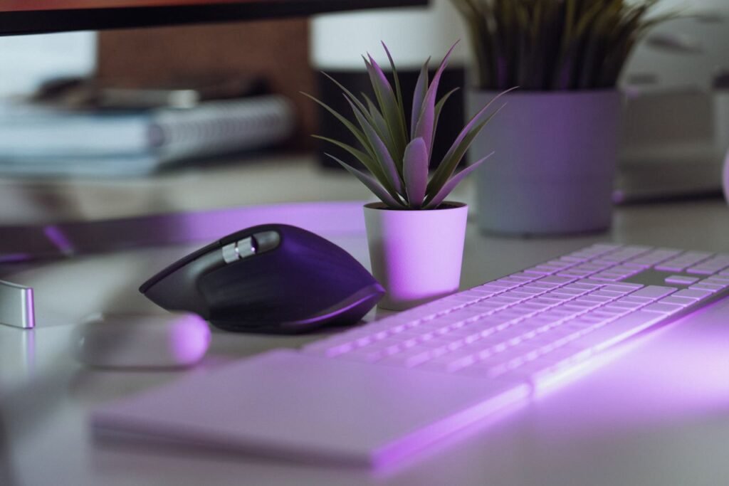 Espaço de escritório em casa estiloso com teclado, mouse e planta em vaso sob iluminação roxa.